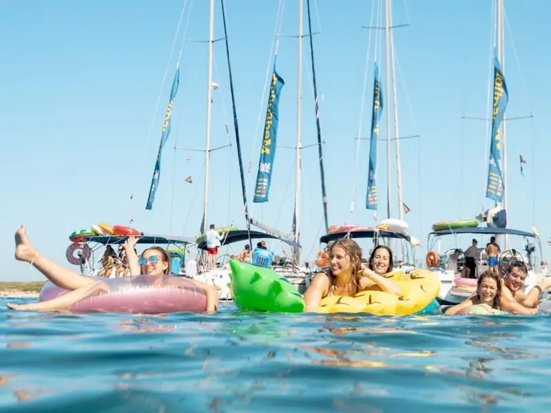 Group of young travellers floating in clear blue water on colourful inflatable rings, laughing and chatting with several sailing yachts anchored behind them under a bright summer sky. The scene captures an easy, social travel vibe in Europe, where you spend the day swimming, meeting new people, and hopping between coastal spots without complicated planning.