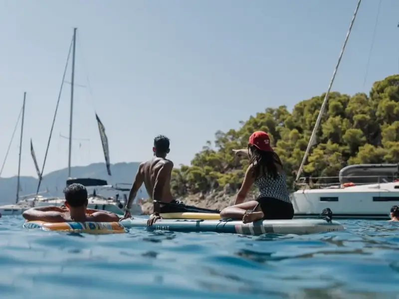 MedSailors guests relaxing on paddleboards in a sheltered bay with sailing yachts anchored nearby and a green hillside behind.