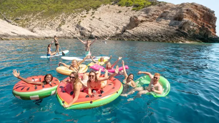 A group of young travelers in their 20s and 30s laughing on colorful watermelon and donut inflatables in the turquoise waters of a secluded bay in Croatia; perfect for those seeking adventure and small group sailing tours in Greece and Croatia for summer 2026.