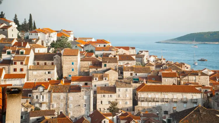 Sunlit view over a coastal European old town with pale stone buildings and terracotta rooftops stacked on a hillside beside a calm blue sea. A few sailboats sit offshore near a green island on the horizon, capturing a warm Mediterranean vibe that feels ideal for a first-time Europe trip with culture, coastline, and relaxed exploring.