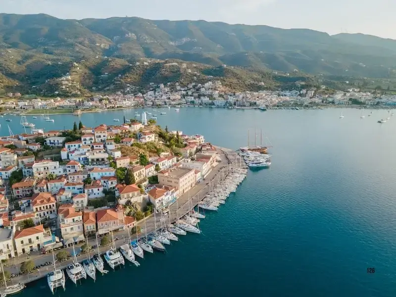 Aerial view of the bustling port town of Poros, Greece, featuring MedSailors yachts moored along the waterfront, historic white-washed buildings with orange-tiled roofs, and lush green mountains overlooking the Saronic Gulf.