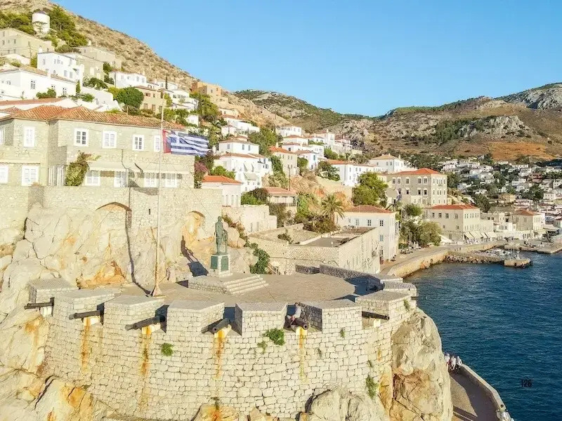 Aerial view of the historic stone-walled port and fortified town of Hydra, Greece, featuring traditional white-washed houses, the Greek flag, and ancient cannons overlooking the deep blue Aegean Sea.