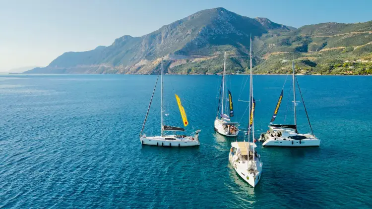 Aerial view of MedSailors sailing yachts and catamarans anchored in the turquoise waters of the Saronic Gulf, Greece, with rugged Mediterranean mountains in the background.