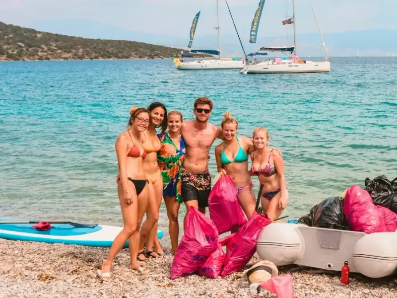 MedSailors guests and crew posing with bags of litter collected during a beach clean on a rocky Mediterranean coastline with sailing yachts anchored in the bay behind them