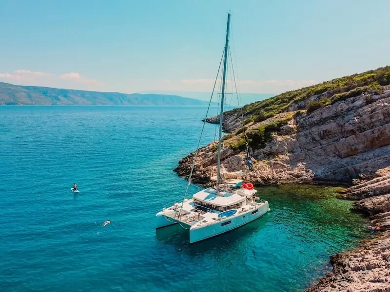 Catamaran anchored in a quiet rocky cove in Croatia with travellers swimming and paddleboarding in clear turquoise water, a classic stop on island hopping and sailing holidays.