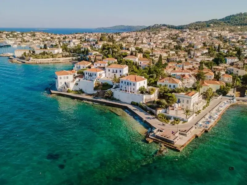 Aerial view of Spetses Island, Greece, showing the turquoise coastline, white-washed seaside villas with orange-tiled roofs, and the winding ring road popular for quad bike and scooter adventures.