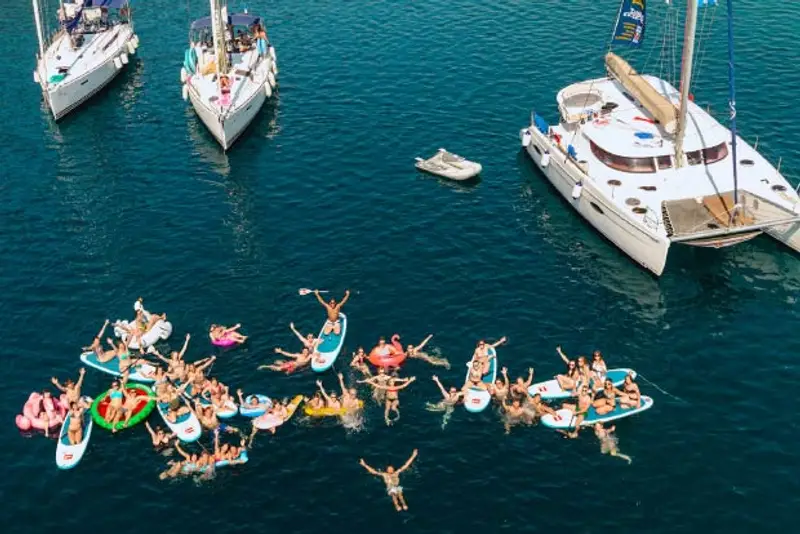 Photo of a large group of people floating on inflatables in turquoise waters of Epidavros Greece with sailing yachts around them.