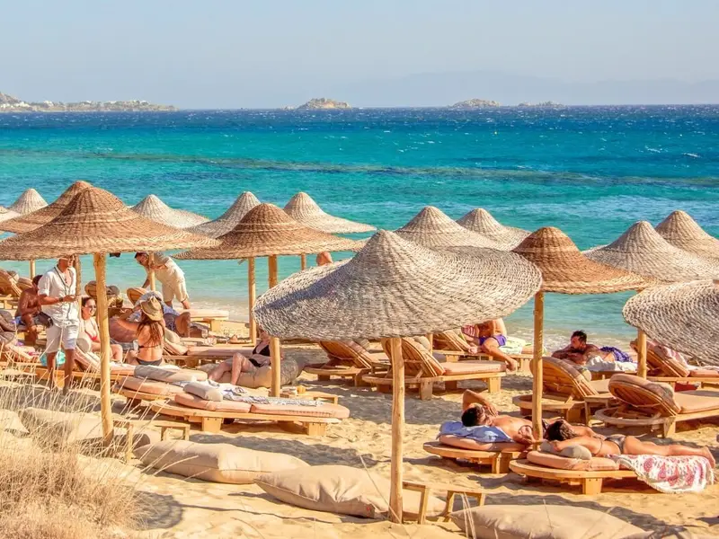Beach scene in Crete, Greece, with straw parasols and wooden sunbeds set on golden sand beside bright turquoise Aegean water, as travellers relax and swim along the coastline. Image used in a MedSailors Odyssey locations in Greece guide to show Crete as a major mythological island in the wider Homeric world, blending ancient legends, coastal landscapes, and modern beach culture into a realistic Greece travel itinerary.