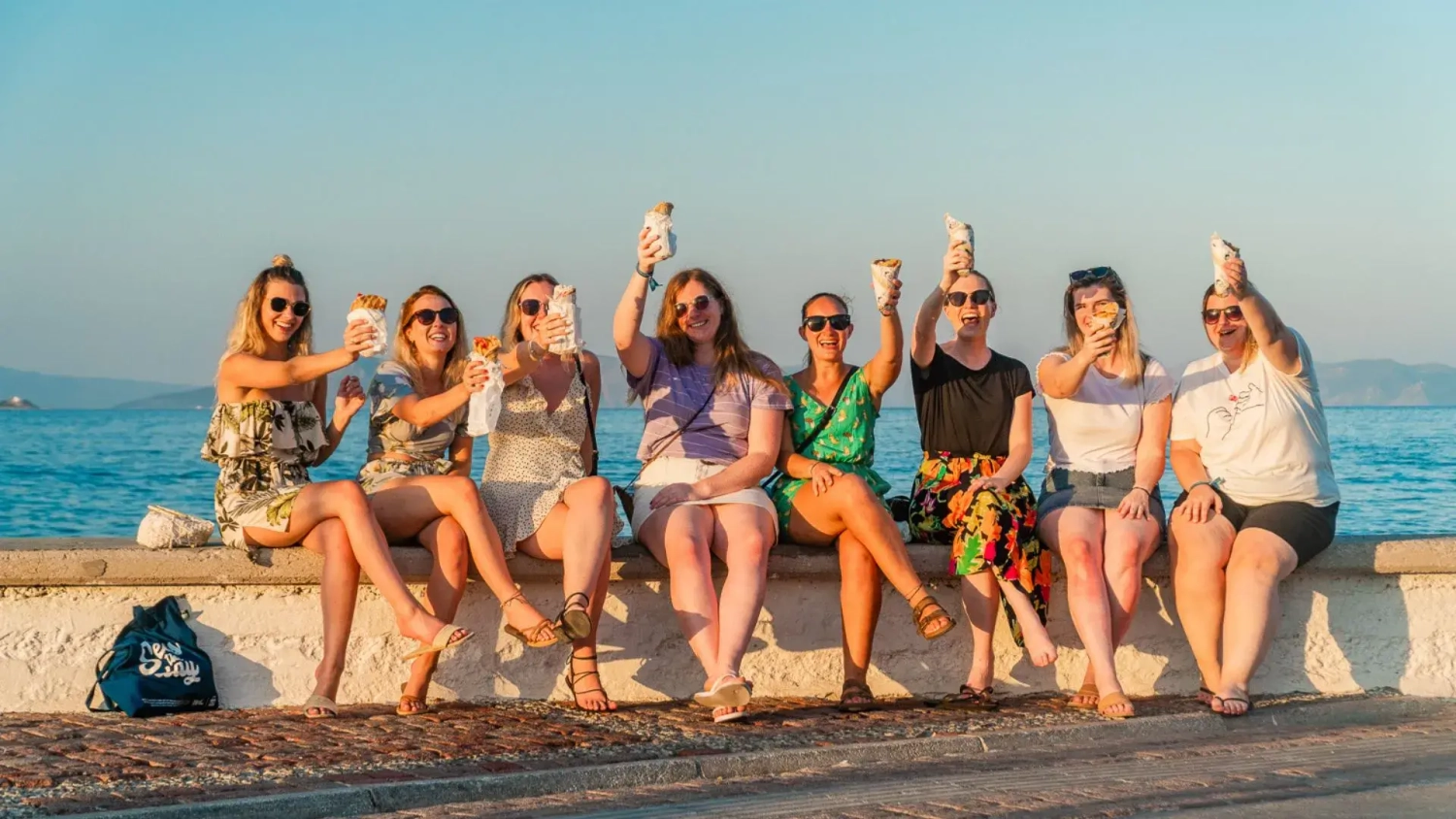 Group of young travellers enjoying Greek street food during sunset on a budget-friendly Greek island hopping trip from Athens. Friends sit by the sea laughing and raising gyros after a day of sailing, swim stops, and exploring local island towns with MedSailors. Social summer travel experience in Greece for solo travellers, couples, and groups seeking affordable island hopping in Europe.