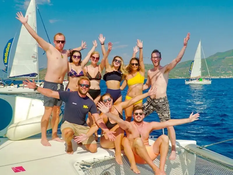 Group of MedSailors travellers posing on a catamaran in the Mediterranean Sea, with crew and yachts in the background during a sailing trip through Greece or Croatia.