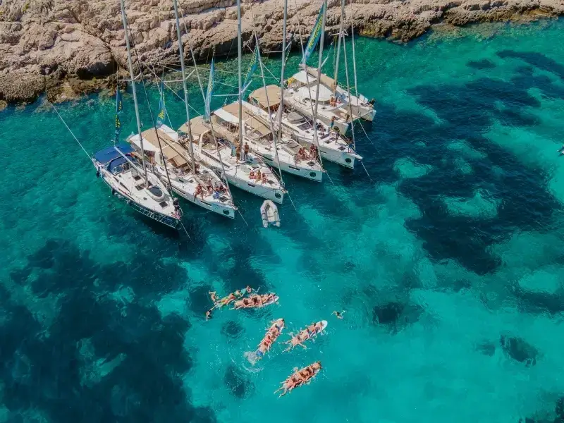 Aerial view of yachts anchored in clear turquoise water along the Croatian coastline, with travellers swimming and relaxing during an island-hopping sailing trip. This image captures the essence of backpacking and group travel in Europe, especially across Croatia and Greece, where sailing, swim stops, and social experiences are central to the journey. Ideal for illustrating Europe travel, island hopping, sailing holidays, and the importance of travel insurance for active trips.