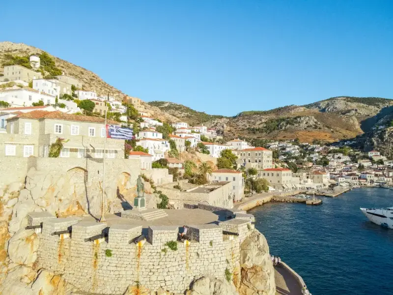 Aerial view of Hydra island harbour with whitewashed houses and stone fort walls during a Greek island hopping trip from Athens. Scenic stop on a budget-friendly summer sailing holiday in Greece, showcasing coastal views, historic architecture, and clear blue Mediterranean waters.