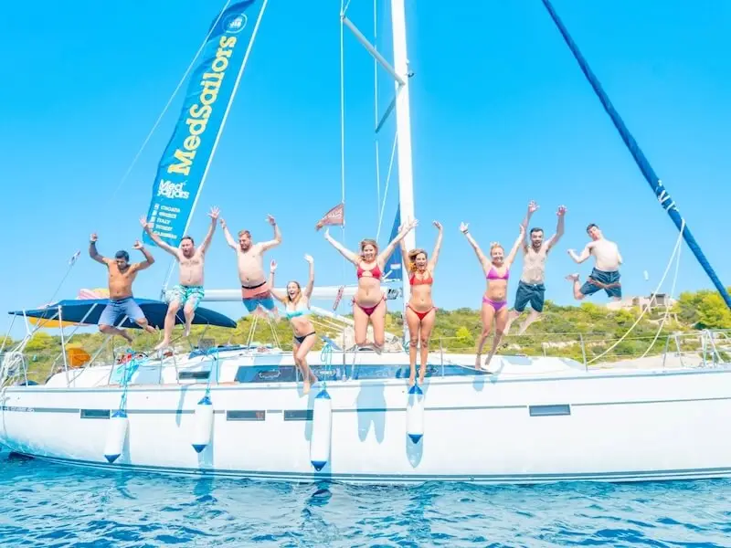 Group of MedSailors travellers jumping off a yacht into the clear blue sea under sunny skies, with the MedSailors sail banner visible — capturing the fun, social energy of a sailing holiday in Croatia or Greece.