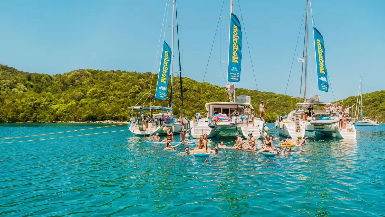 MedSailors sailing yachts at anchor on a raft with a group of young 20s and 30s travellers floating in a bright turquoise bay in the Mediterranean on their social island hopping sailing adventure in Greece and Croatia.