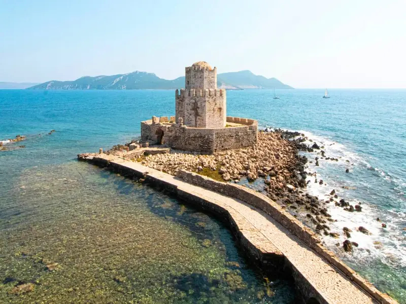 Aerial view of Methoni Castle near Pylos in the Messenia region of the Peloponnese, Greece, with a stone causeway leading across shallow clear water to a fortified coastal tower and walls surrounded by the sea. Image used in a MedSailors Odyssey locations in Greece guide, highlighting Pylos as a key stop in Homer’s world where Telemachus visits Nestor during The Odyssey, and linking the area’s historic coastal sites with a modern Greece travel itinerary.