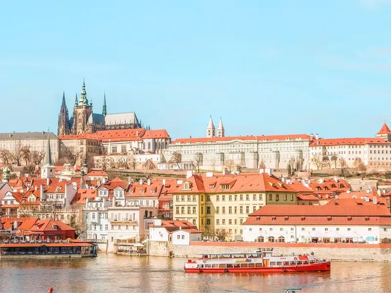 Panoramic view across Prague’s red-roofed old town with Prague Castle and St Vitus Cathedral rising on the hill in the background, seen from the Vltava River with a small boat passing in the foreground. The scene captures Prague’s fairytale architecture, walkable historic centre, and classic riverside viewpoints, ideal for first-time Europe travellers looking for culture, scenery, and good value.