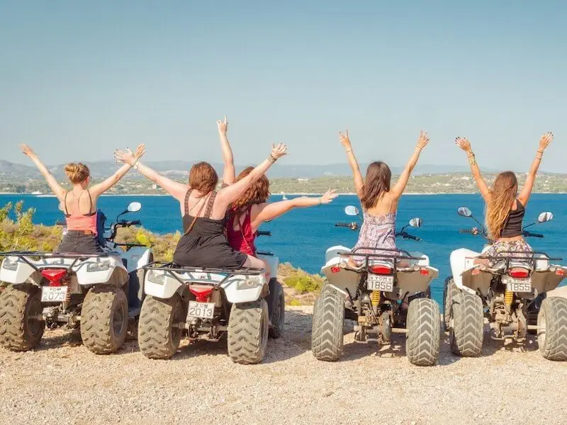 Group of MedSailors travellers on quad bikes with arms raised, overlooking the sea and coastline in Greece during an island-hopping adventure.