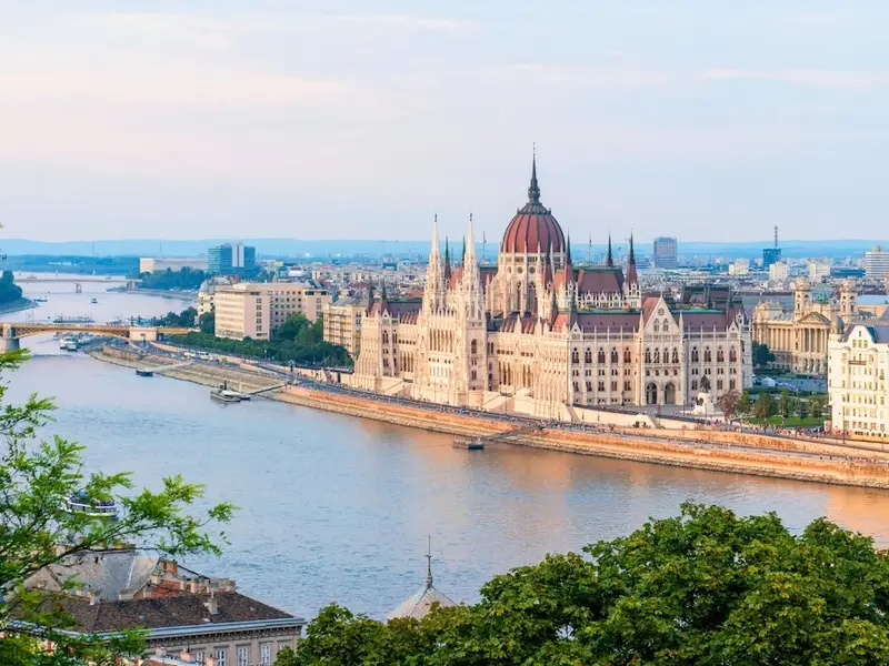Panoramic view of Budapest’s Hungarian Parliament Building glowing beside the Danube River, seen from across the water with bridges and city skyline stretching into the distance. The scene captures Budapest’s grand architecture, riverside walking routes, and classic city-break atmosphere, ideal for first-time Europe travellers looking for culture, views, and great value.