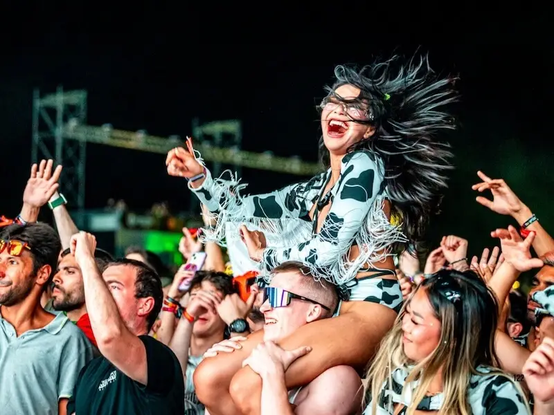 A happy woman in a cow-print festival outfit laughing on a friend's shoulders at a 2026 European summer music festival during a MedSailors island hopping trip.