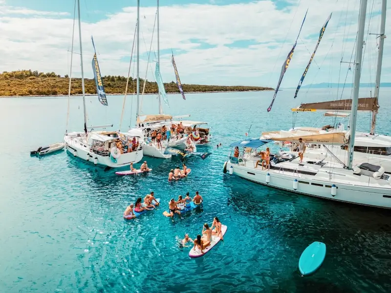 Group of MedSailors yachts rafted together in turquoise water, with travellers swimming, paddleboarding, and relaxing between boats — showcasing the fun and social atmosphere of a MedSailors sailing holiday in Croatia or Greece.