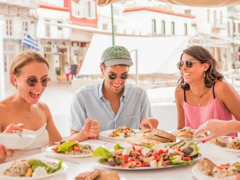 Group of smiling budget travellers sitting in front of Greek food in a tradtional town while island hopping Greece with MedSailors.