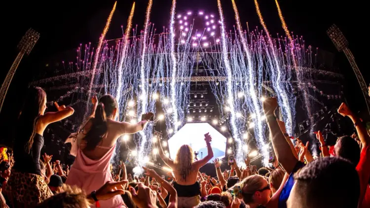 A vibrant night scene at a large outdoor music festival in Europe with a massive stage, bright fireworks shooting into the sky, and a cheering crowd with their hands raised in the air. Part of a group tour for 18-35 travellers combining ULTRA festival with Croatia island hopping on MedSailors.