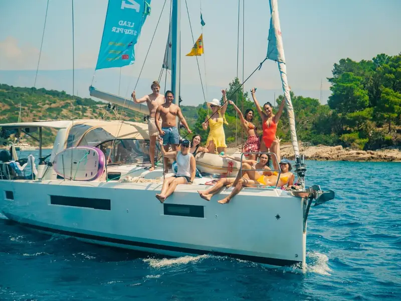 A diverse group of young travelers smiling and cheering on the deck of a white sailing yacht in a lush, pine-fringed bay in Greece; perfect for 18-35s looking for social group travel and Mediterranean adventure holidays.