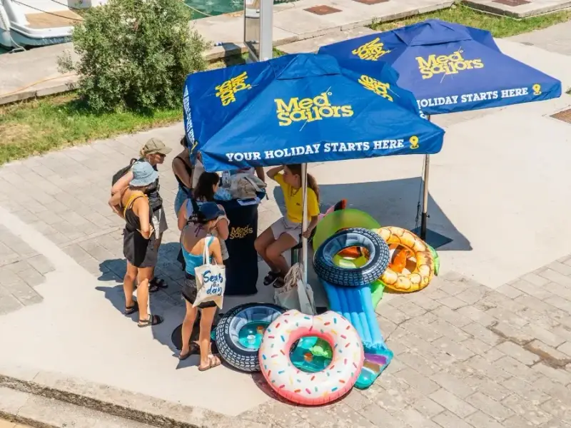 MedSailors check-in desk at a Mediterranean port with colourful inflatables ready for floatie recycling and a guest carrying a MedSailors tote bag.