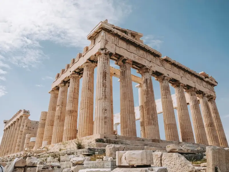 Upward view of the Parthenon on the Acropolis in Athens, showing tall ancient marble columns and weathered stone blocks under a pale blue sky. The scene highlights Athens’ iconic classical architecture and must-see ancient history, ideal for first-time Europe travellers planning an easy city break with major landmarks, walkable neighbourhoods, and rooftop views nearby.
