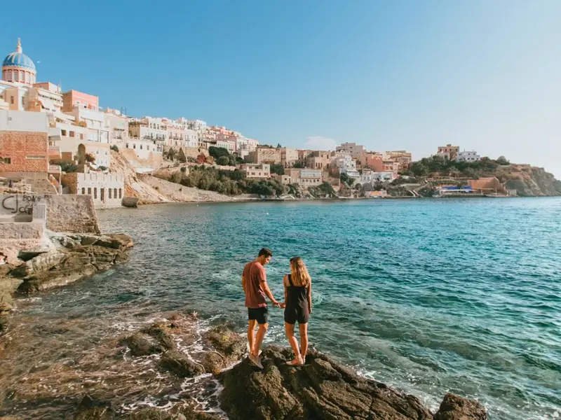Couple standing on rocky shoreline looking across the sea toward Ermoupoli, the main town of Syros in the Cyclades, with pastel buildings and a domed church rising above the waterfront under a clear blue sky. Image used in a MedSailors Odyssey locations in Greece guide to support the Cyclades section, linking Syros with Homer’s “Syrie” tradition and showing a real, visitable island base for an Odyssey-inspired Greece itinerary.