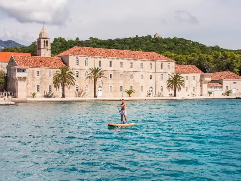 Person stand-up paddleboarding on clear blue water in front of a historic coastal town in Croatia.