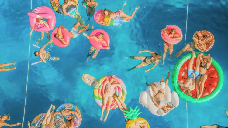 Aerial view of MedSailors travellers swimming and relaxing on colourful inflatable floats in crystal-clear Mediterranean waters during a group sailing holiday, showcasing the fun and social atmosphere of island hopping with friends.