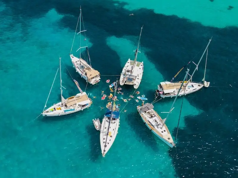 Aerial photo of a MedSailors sailing holiday flotilla of yachts anchored together in crystal-clear turquoise water, with travellers swimming, paddleboarding, and relaxing between boats — capturing the social sail week in Croatia or Greece.