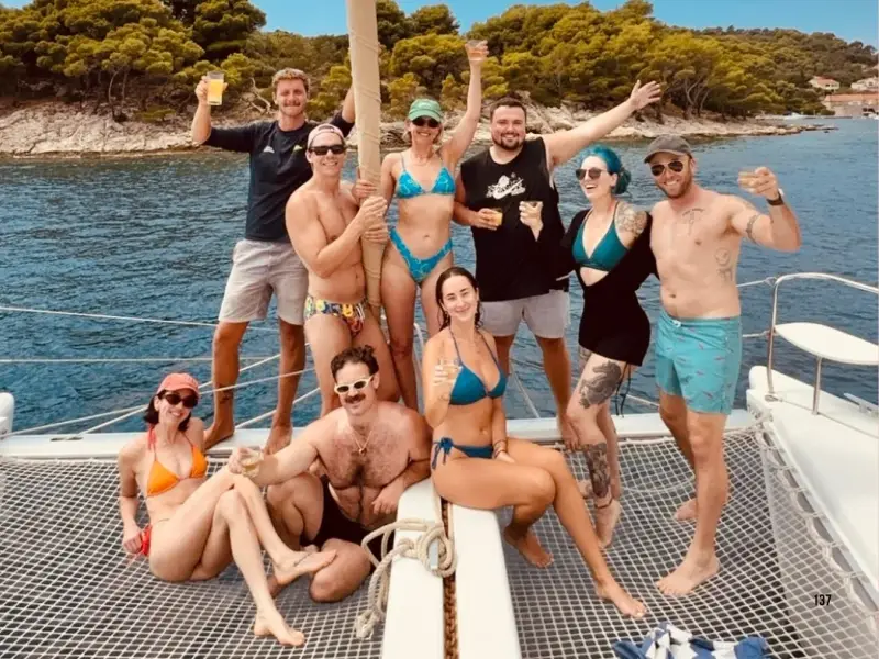 Group of young travellers celebrating on a catamaran during a Croatia island-hopping trip, with clear Adriatic water and pine-covered coastline in the background. A great image for a guide to ULTRA Europe and how to turn the festival into a full Croatia sailing and summer travel experience.