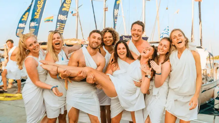 MedSailors travellers laughing and celebrating together in white outfits beside a flotilla of sailing yachts, capturing the friendships and social atmosphere of a Mediterranean group sailing holiday.