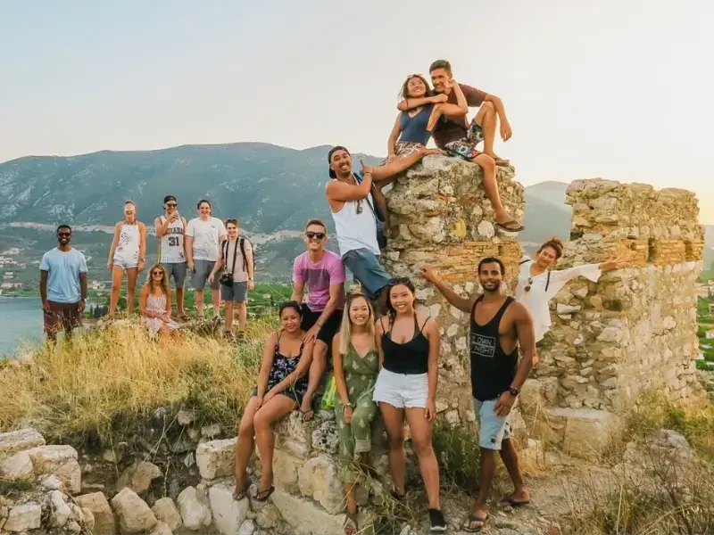 Group of young backpackers and travellers exploring historic ruins in Greece with scenic mountain views in the background, capturing the social and adventurous side of a Europe trip. This image reflects group travel experiences across Croatia and Greece, where travellers explore cultural landmarks, hike to viewpoints, and share moments with new friends. Ideal for illustrating backpacking Europe, island-hopping travel, and planning a social summer trip with travel insurance for peace of mind.