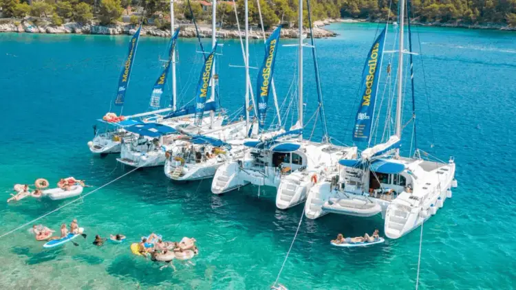 Aerial view of MedSailors sailing yachts and catamarans rafted together in a turquoise Mediterranean bay, with travellers swimming, paddleboarding and relaxing on inflatables during a social sailing holiday.