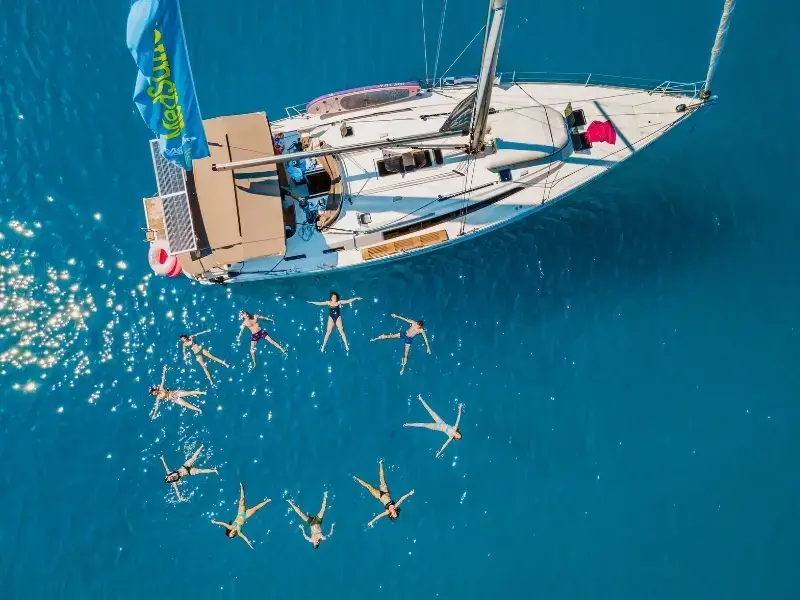 Aerial view of backpackers and young travellers floating in the sea beside a yacht during a swim stop on a Europe island-hopping trip in Croatia. This scene captures the relaxed and social nature of sailing holidays, where travellers explore hidden bays and spend time in the water, making it ideal for illustrating backpacking Europe, sailing trips, and travel planning for active group adventures.