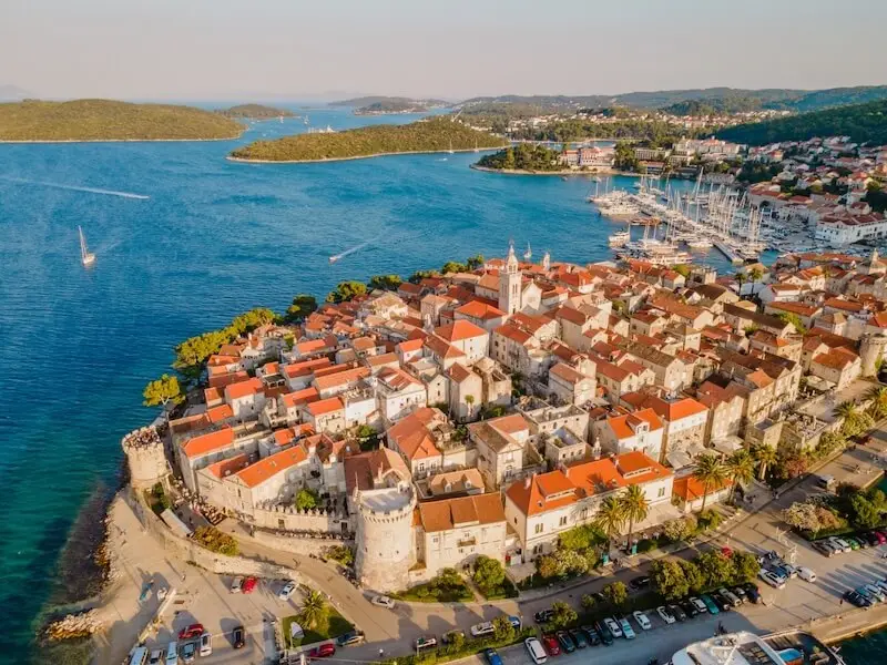Aerial view of Korčula Old Town, Croatia, showing its medieval stone walls, red-roofed houses, and surrounding turquoise sea with sailboats anchored nearby — a highlight destination on MedSailors’ Croatia sailing holidays.