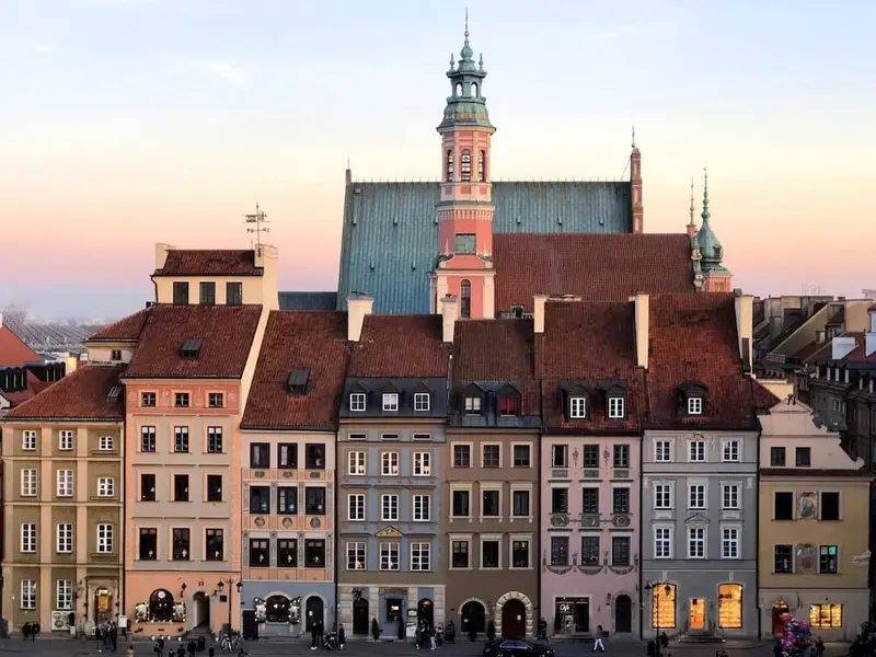 Sunset view of colourful historic townhouses in Warsaw’s Old Town, with pastel façades and red-tiled roofs lining a lively square and a tall church tower rising behind them. Warm evening light hits the rooftops as people gather below, capturing Warsaw’s walkable, first-timer friendly atmosphere and classic Central European architecture on a budget-friendly city break.