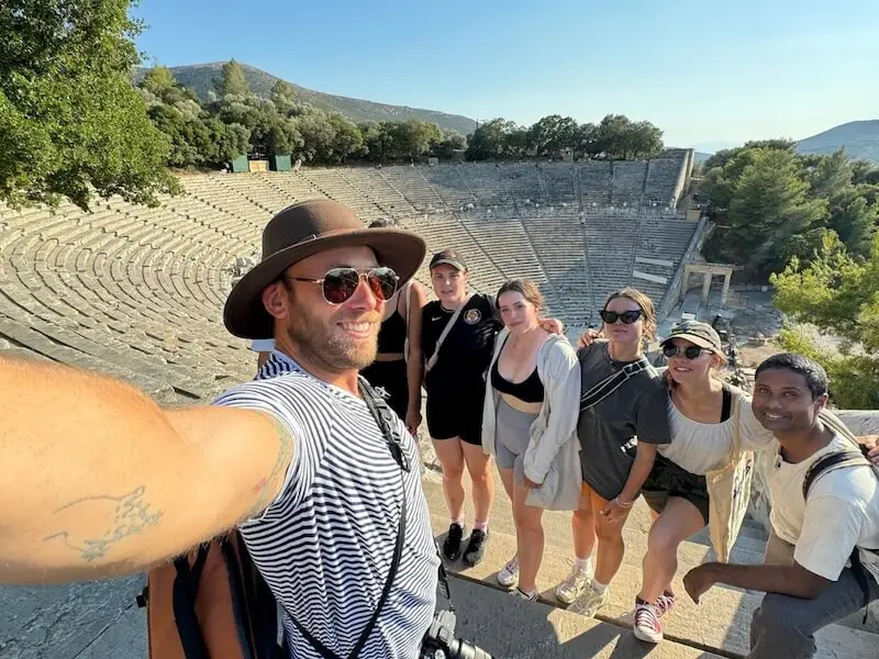 Group of MedSailors travellers taking a selfie at the Ancient Theatre of Epidaurus in Greece, with the historic stone amphitheatre in the background.