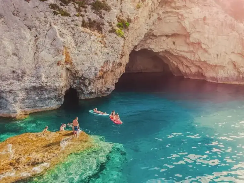 Travellers swimming and paddleboarding in clear blue water beside a rocky cove and a large sea cave entrance, with warm sunlight flaring across the cliffs. Photo supports a MedSailors Odyssey locations guide, illustrating the best parts of a modern Odyssey-style trip in Greece: hidden coves, sea caves, swim stops, and sailing holiday adventure experiences.