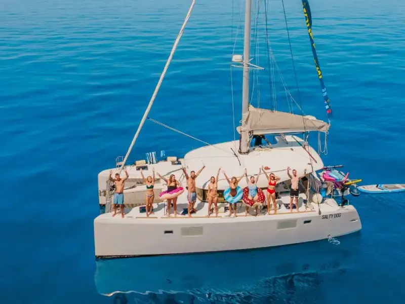 Aerial photo of MedSailors guests and crew standing on a catamaran deck with arms raised above calm blue water.