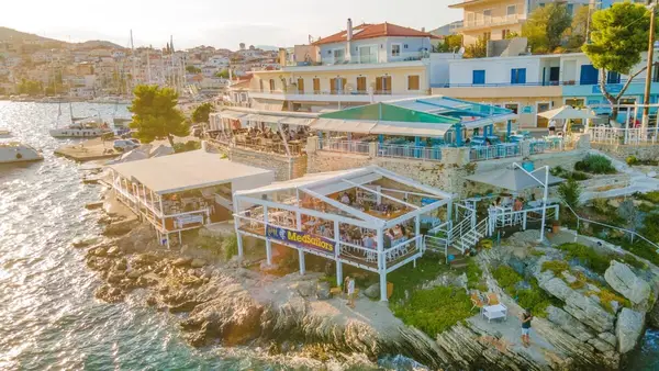 Ermioni town aerial at sunset showing seaside cocktail bars and restaurants along the coastline of Ermioni town harbour, one of the best food scenes in Greece.