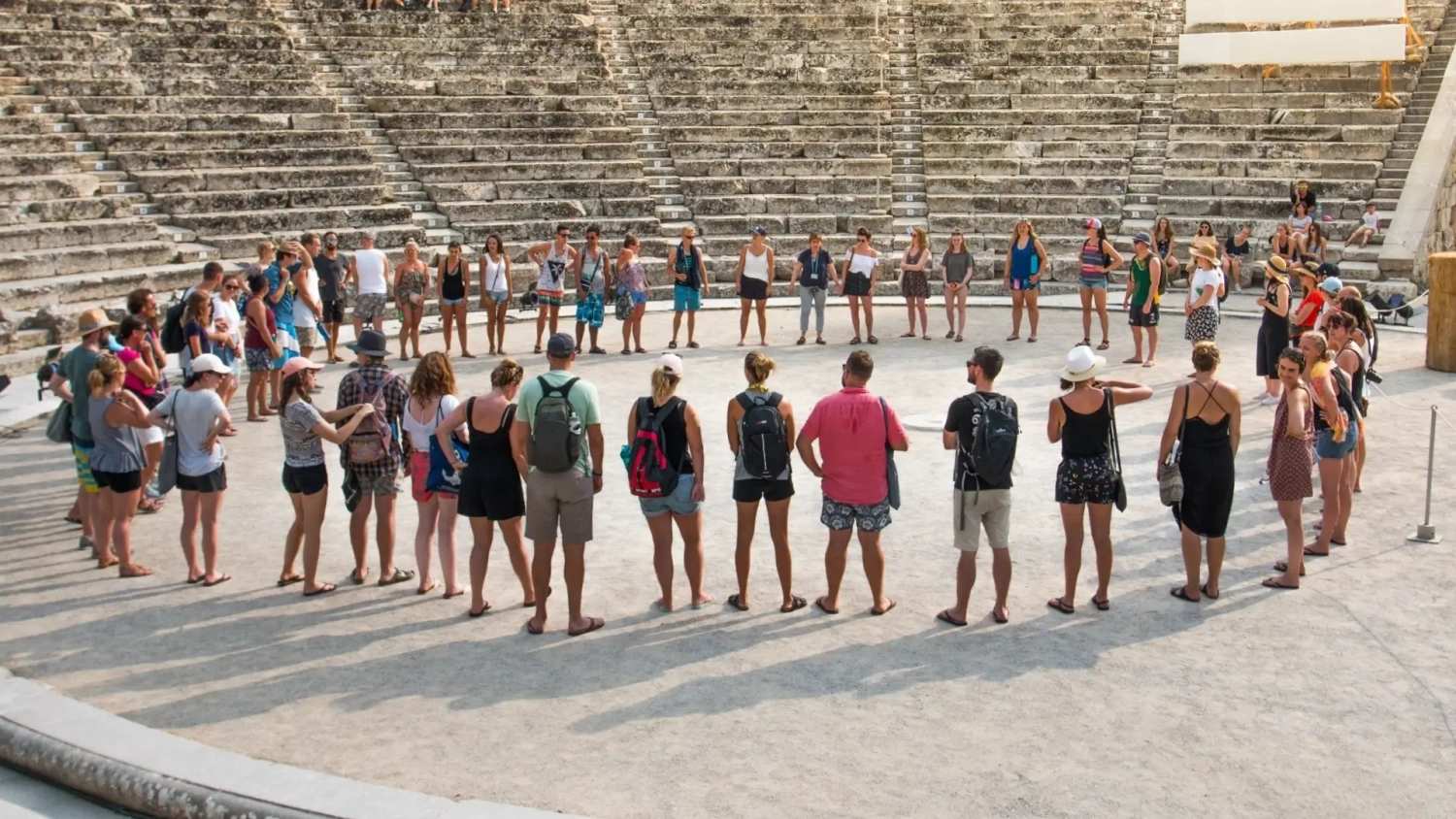 Group of young backpackers and travellers standing in a circle inside an ancient theatre in Greece during a guided tour, capturing the social and cultural side of a Europe trip. This scene reflects the kind of group travel experiences common on island-hopping routes across Croatia and Greece, where history, community, and shared moments are a key part of the journey. Ideal for illustrating Europe backpacking trips, travel insurance for young travellers, and group adventure travel experiences.