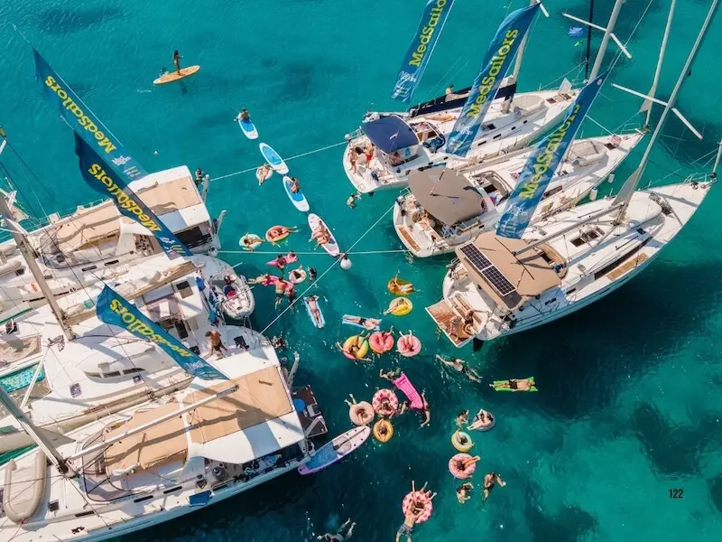 Aerial view of MedSailors yachts rafted together in turquoise Mediterranean waters during a 2026 Europe festival group tour, with guests swimming and using paddleboards.