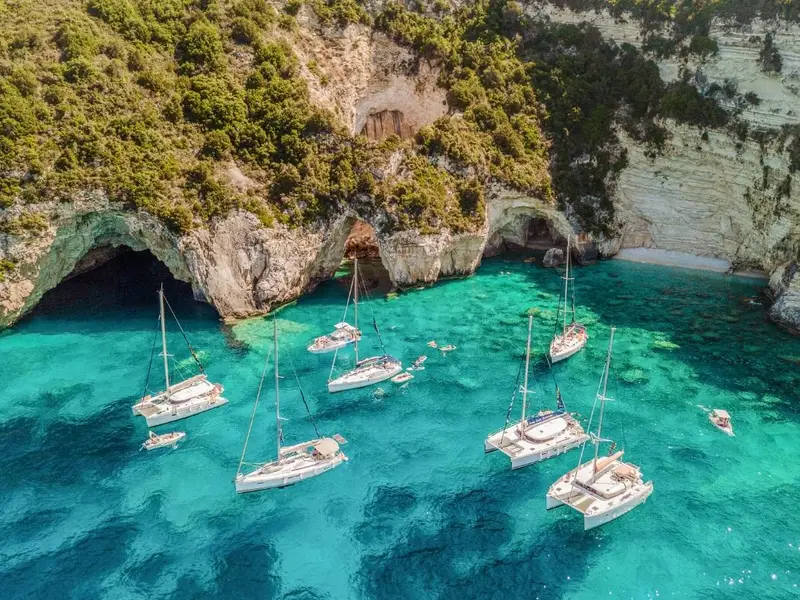 Aerial view of multiple sailing yachts anchored in clear turquoise water beneath dramatic limestone cliffs and large sea caves in Corfu, Greece. Swimmers float between the boats in a sheltered Ionian cove. Image used in a MedSailors guide to Odyssey locations you can visit in Greece, linking Corfu with Scheria, the land of the Phaeacians in Homer’s The Odyssey, and showcasing island hopping, sailing holidays, and Ionian coastline adventure travel.