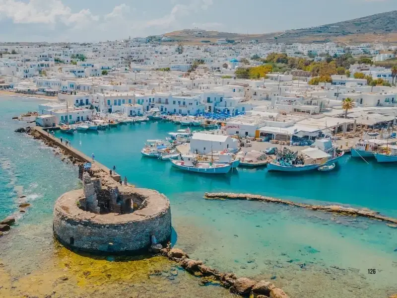A vibrant aerial view of Paros Island's charming harbour in the Cyclades, with whitewashed buildings, colourful fishing boats, and turquoise waters – showcasing the relaxed beauty and authentic culture that make Greek island hopping unforgettable.