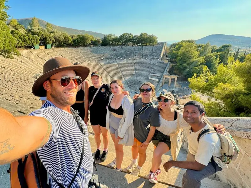 Group of travellers taking a selfie at the ancient theatre of Epidaurus (Epidavros) in the Peloponnese, Greece, with the huge stone amphitheatre seating rising behind them in warm late-afternoon light. Image used in a MedSailors Odyssey locations in Greece guide, linking real historic sites near Athens to Homer’s wider world and the Trojan War era, and showing how a modern Greece itinerary can mix island hopping, archaeology, and day trips like Epidaurus.
