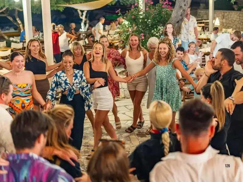 A lively group of young travellers joins hands in a circle, dancing at a traditional Greek taverna under fairy lights — celebrating the spirit of island life and local culture during a MedSailors sailing adventure.